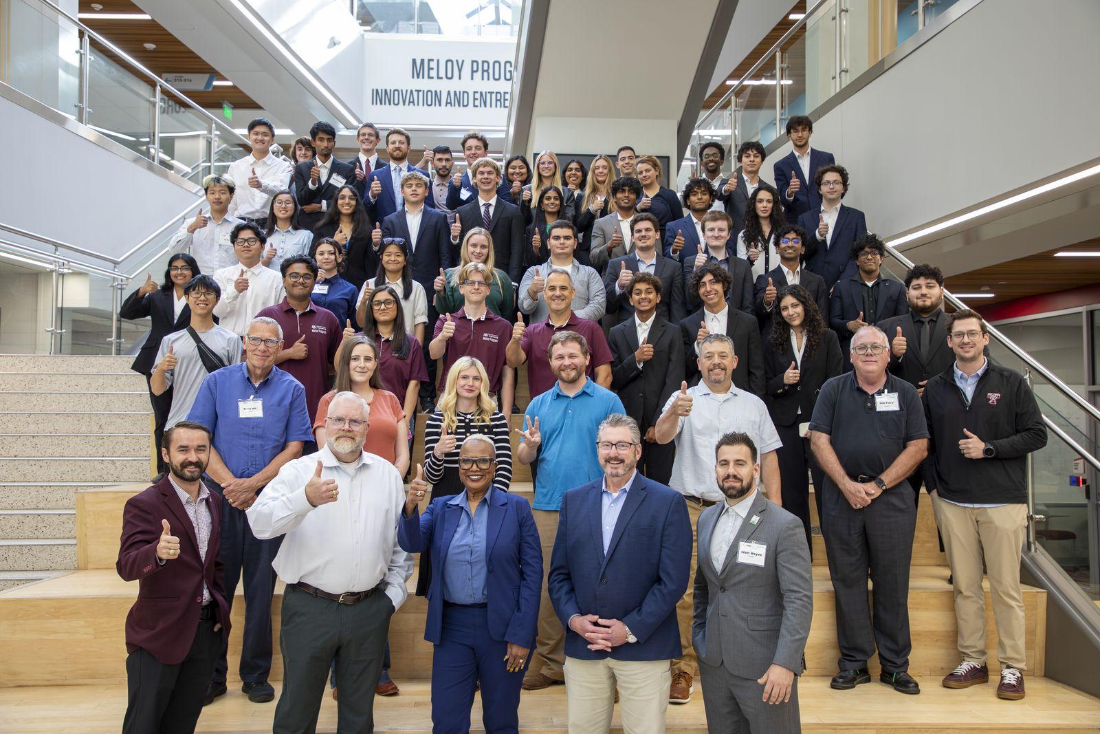Aggies Invent students, mentors and judges giving a thumbs up on the stairs under the Meloy Program sign