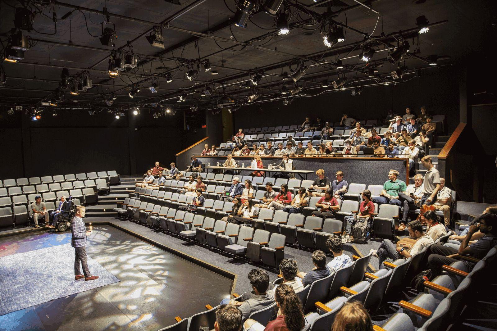 A presenter on stage in a black-box theater, with judges and an audience of students in the seats
